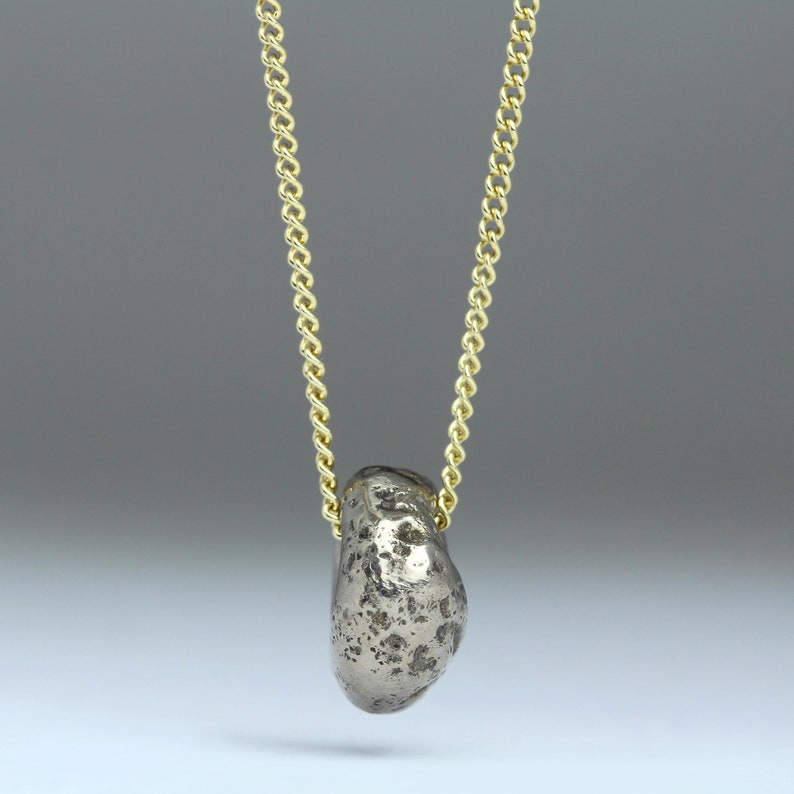 Puede incluir: Un collar con un colgante de plata de forma de roca, texturizado y rugoso, que cuelga de una cadena dorada.