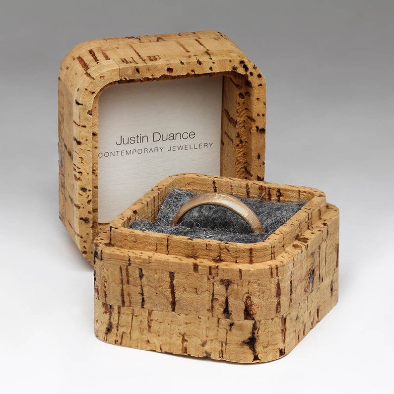 May include: A gold ring sits in an open, square cork jewellery box with a grey interior. The box displays the ring and a white card with the text "Justin Duance CONTEMPORARY JEWELLERY". The box has a natural, textured appearance.