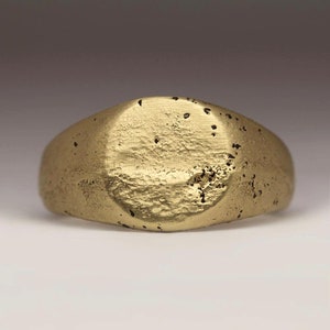 May include: A gold signet ring with a smooth, oval-shaped top. The ring has a textured surface.