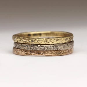 May include: Three thin gold, silver, and rose gold bands with a textured finish. The bands are stacked on top of each other.