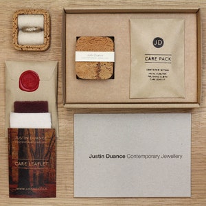 May include: A brown cardboard box with a cork coaster inside. The box also contains a brown paper envelope with a wax seal, a small brown paper bag with the text "CARE PACK" and the text "CONTAINED WITHIN: METAL SCOURER, POLISHING CLOTH, CARE LEAFLET", and a white card with the text "Justin Duance Contemporary Jewellery".