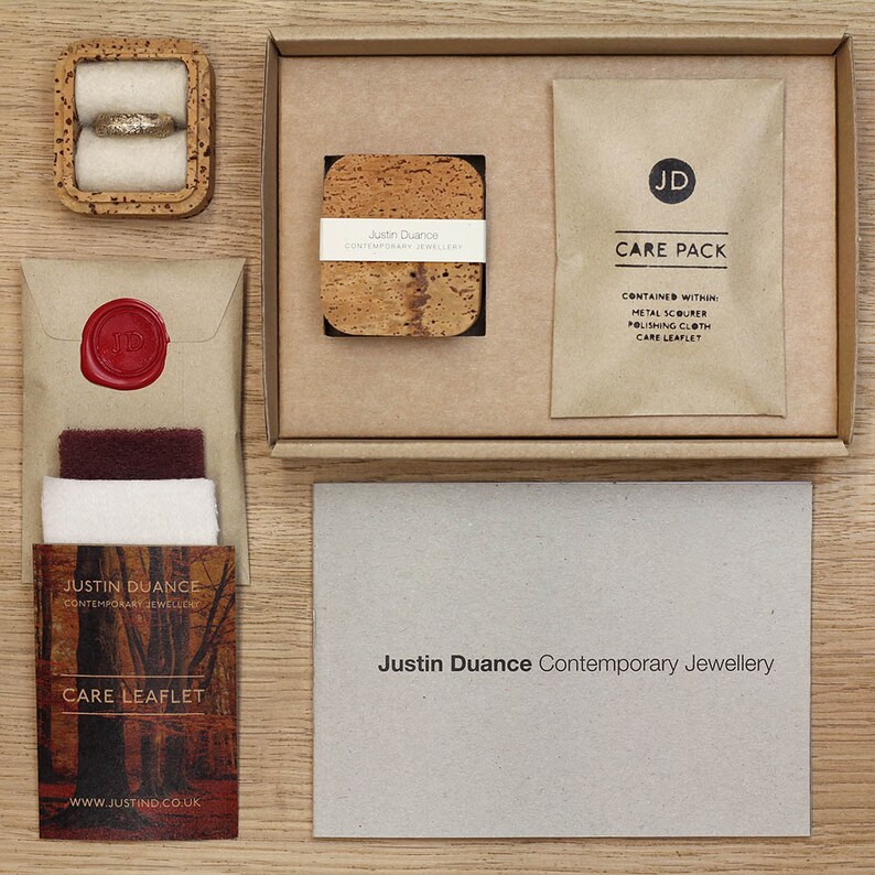May include: A brown cardboard box with a cork coaster inside. The box also contains a brown paper envelope with a wax seal, a small brown paper bag with the text "CARE PACK" and the text "CONTAINED WITHIN: METAL SCOURER, POLISHING CLOTH, CARE LEAFLET", and a white card with the text "Justin Duance Contemporary Jewellery".