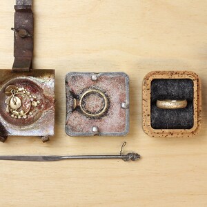 May include: A close-up of a jewelry making process, showing a small jar of sand, a metal mold with a gold pendant, a metal mold with a gold ring, a cork box with a gold ring, and a cork box with the text "Justin Duance Contemporary Jewellery".