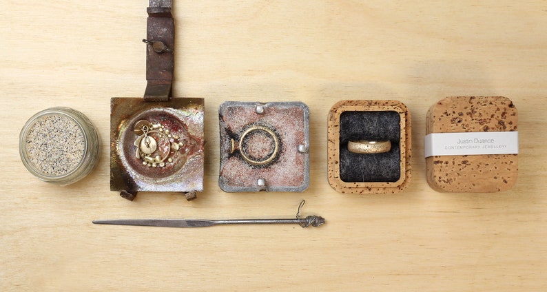 May include: A close-up of a jewelry making process, showing a small jar of sand, a metal mold with a gold pendant, a metal mold with a gold ring, a cork box with a gold ring, and a cork box with the text "Justin Duance Contemporary Jewellery".