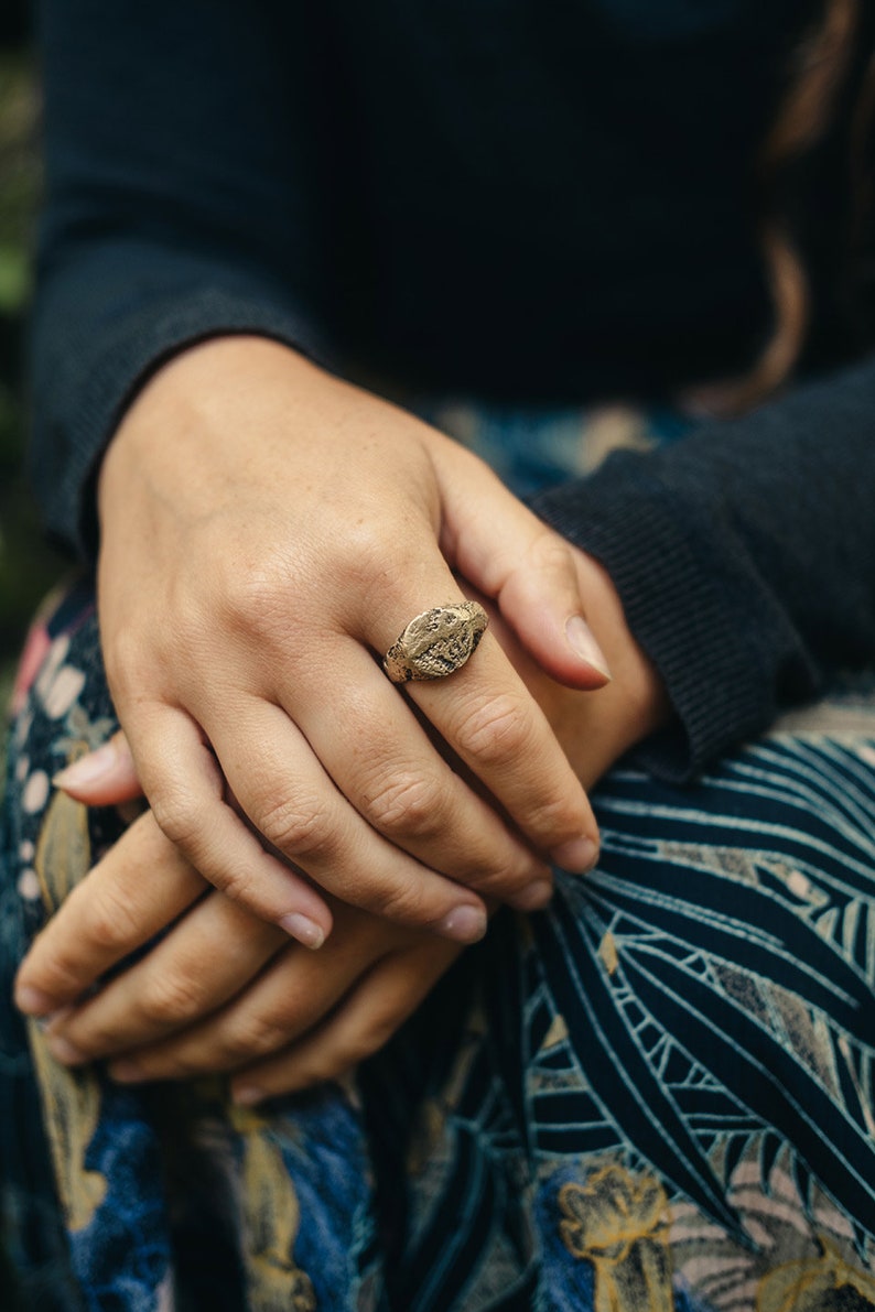 May include: A gold ring with a detailed design on the top. The ring is worn on the finger of a person wearing a dark blue shirt and a patterned skirt.