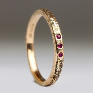 May include: A gold ring featuring a textured finish and three small, round, red gemstones. The ring's band is simple, and the overall appearance suggests a handcrafted or vintage design. The gemstones are set in a row.