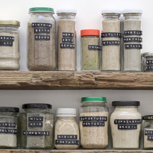 May include: A display of glass jars filled with sand from various locations, each jar labelled with a place name. The jars are arranged on rustic wooden shelves. Labels include "Devon," "Ireland," and "Dorset."