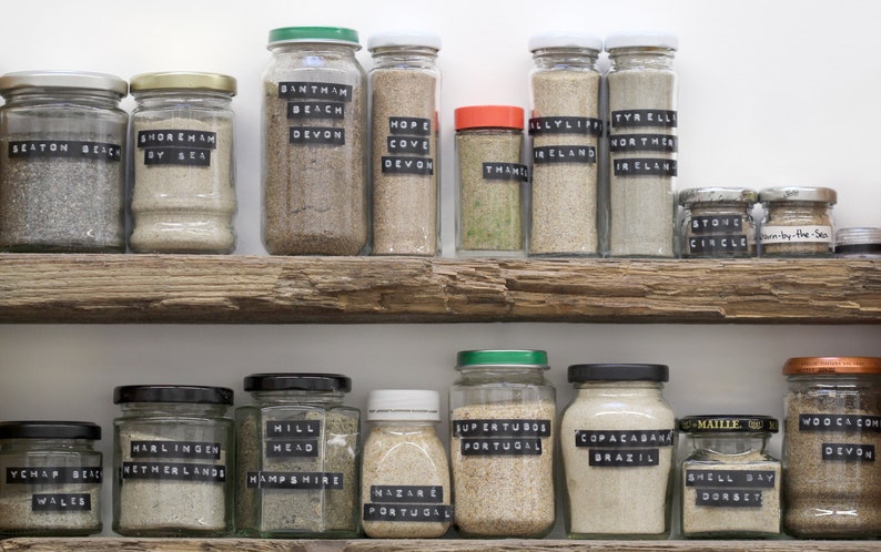 May include: A display of glass jars filled with sand from various locations, each jar labelled with a place name. The jars are arranged on rustic wooden shelves. Labels include "Devon," "Ireland," and "Dorset."