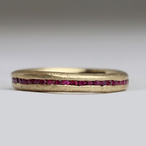 May include: A gold band ring with a single row of small red ruby stones set into the band.