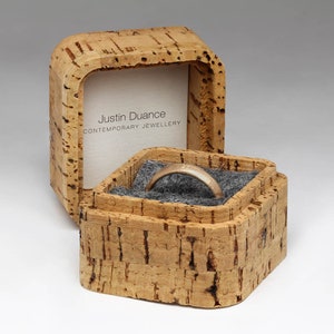 May include: A square cork ring box with a gray felt interior. The box is open and a gold ring is inside. The box has the text "Justin Duance CONTEMPORARY JEWELLERY" on the side.