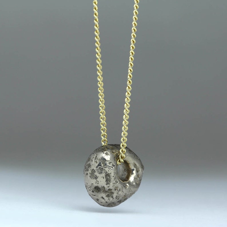 Puede incluir: Un collar con un colgante redondo y hueco de plata texturizado. El colgante est&aacute; unido a una cadena de oro.
