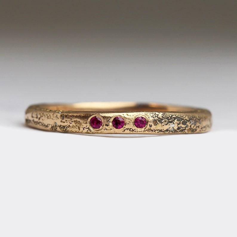 May include: A gold ring featuring three small, round, red gemstones. The ring has a textured surface. The gemstones are set in a line, adding a vibrant contrast to the gold. Jewellery.