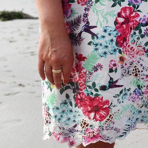 May include: A woman's hand wearing a gold ring, with a floral print skirt and white lace trim. The skirt has a tropical floral pattern with hummingbirds.