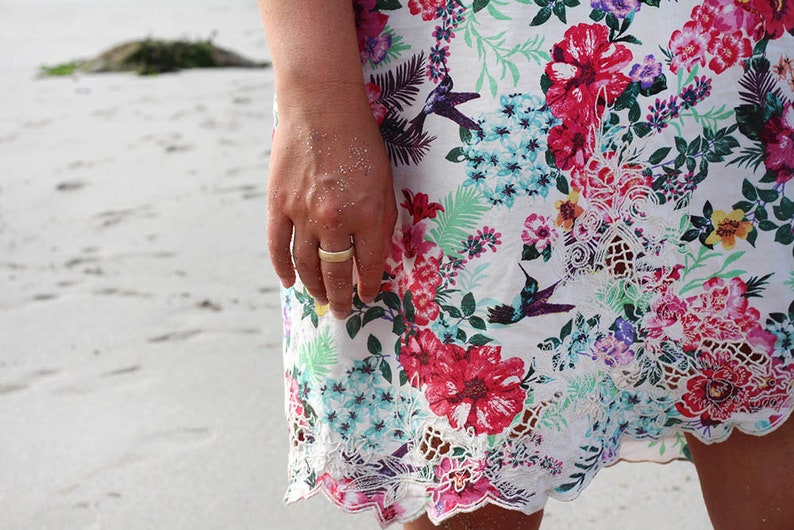 May include: A woman's hand wearing a gold ring, with a floral print skirt and white lace trim. The skirt has a tropical floral pattern with hummingbirds.