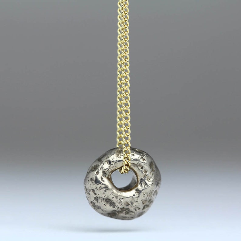 Puede incluir: Un colgante de anillo de plata con una superficie texturizada cuelga de una cadena de oro. El anillo est&aacute; abierto y tiene un agujero en el centro.