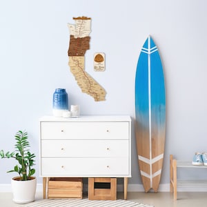 May include: A white dresser with three drawers and gold knobs sits in front of a light blue wall. A wooden surfboard with blue and white accents leans against the wall. A wooden map of California hangs on the wall above the dresser. A potted plant sits on the floor next to the dresser.