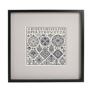 May include: Framed cross-stitch sampler with a black frame. The sampler features a cream-colored background with dark blue cross-stitch designs, including skulls, floral patterns, and geometric shapes. The top of the sampler displays the alphabet.