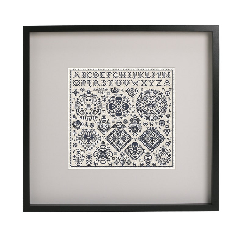 May include: Framed cross-stitch sampler with a black frame. The sampler features a cream-colored background with dark blue cross-stitch designs, including skulls, floral patterns, and geometric shapes. The top of the sampler displays the alphabet.