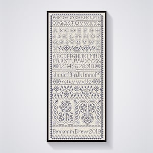 May include: A framed cross-stitch sampler with a white background and blue stitching. The sampler includes the alphabet in uppercase and lowercase, numbers, and a decorative border. The text "Benjamin Drew 2019" is stitched at the bottom.