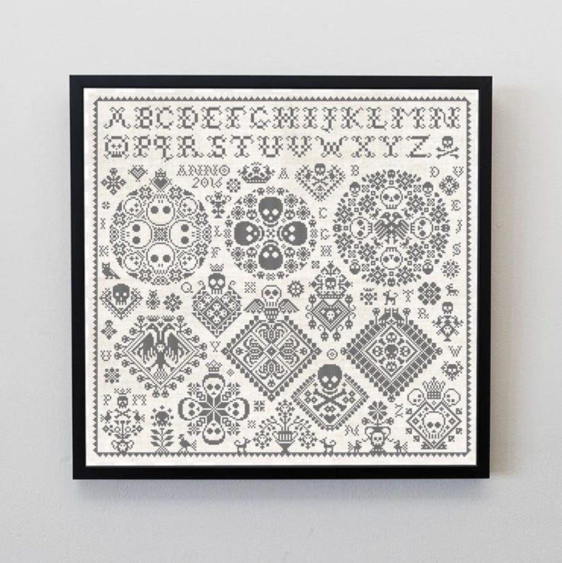 May include: Framed cross-stitch sampler with a black frame. The design features skulls, crowns, and geometric patterns in gray and white. The alphabet and the year 2016 are also included in the design. The overall theme is gothic and macabre.