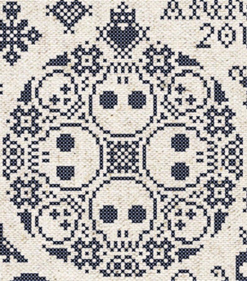 May include: Close-up of a cross-stitch design featuring four skull motifs in a circular pattern. The skulls are surrounded by floral and geometric elements, all in dark blue thread on a cream-colored fabric. The design has a folk art aesthetic.