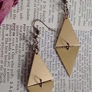 May include: Geometric gold-colored earrings. These earrings feature a unique design with two connected diamond shapes. The earrings are lightweight and suitable for everyday wear. The earrings are approximately 1.5 inches in length.