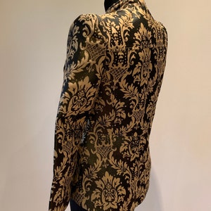 Pode incluir: Um blazer estruturado com gola alta e mangas compridas, decorado com um padr&atilde;o de damasco dourado repetitivo sobre um fundo preto profundo. A pe&ccedil;a &eacute; exibida num manequim, destacando os intrincados motivos florais e o corte ajustado.