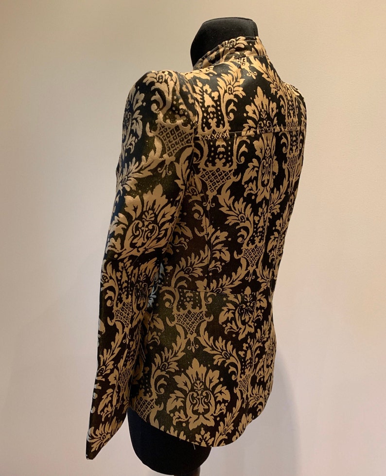 Pode incluir: Um blazer estruturado com gola alta e mangas compridas, decorado com um padr&atilde;o de damasco dourado repetitivo sobre um fundo preto profundo. A pe&ccedil;a &eacute; exibida num manequim, destacando os intrincados motivos florais e o corte ajustado.