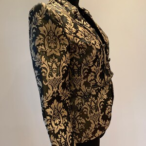 Pode incluir: Um blazer estruturado com um padr&atilde;o de damasco em preto e dourado. A pe&ccedil;a tem gola alta, mangas compridas e fecho frontal com bot&otilde;es met&aacute;licos. O tecido apresenta um motivo floral ornamental repetido por toda a superf&iacute;cie.