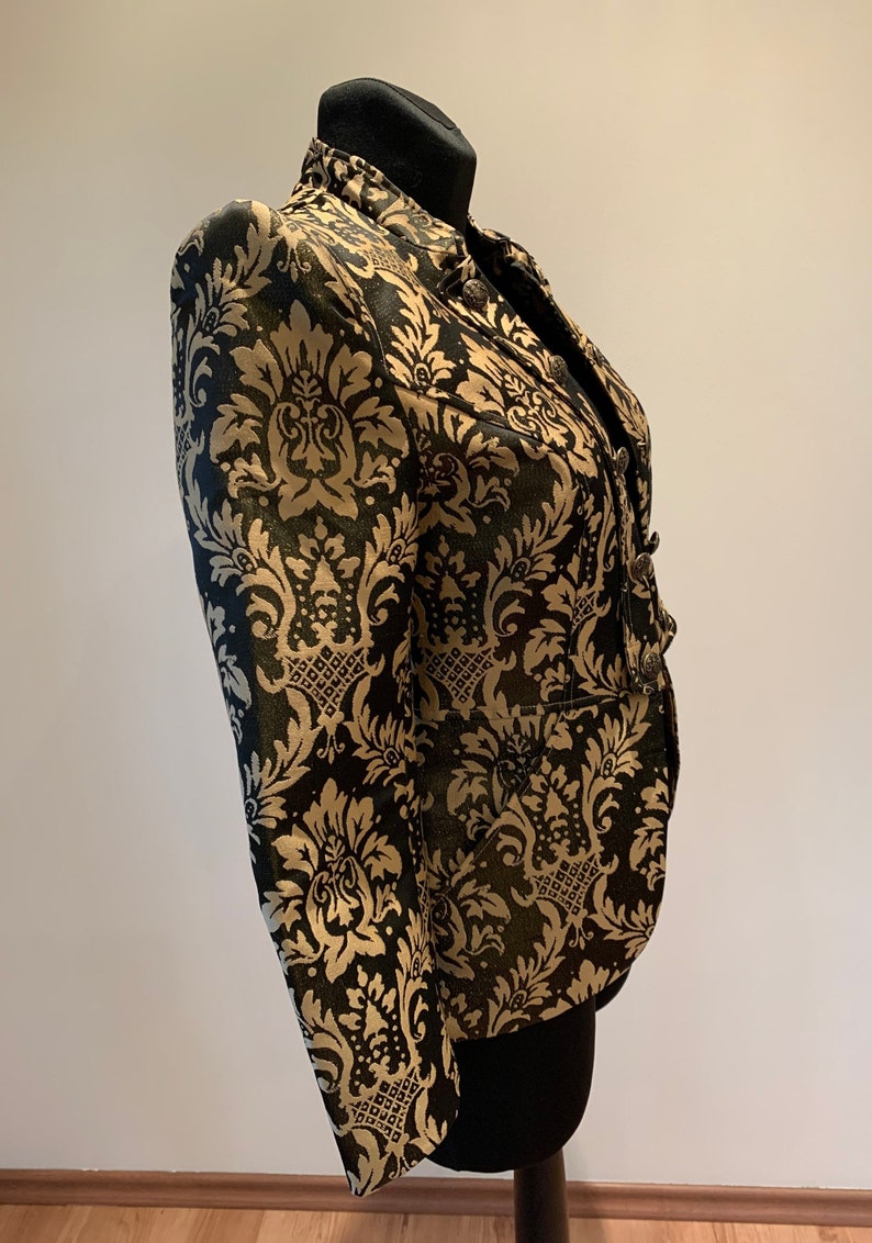 Pode incluir: Um blazer estruturado com um padr&atilde;o de damasco em preto e dourado. A pe&ccedil;a tem gola alta, mangas compridas e fecho frontal com bot&otilde;es met&aacute;licos. O tecido apresenta um motivo floral ornamental repetido por toda a superf&iacute;cie.