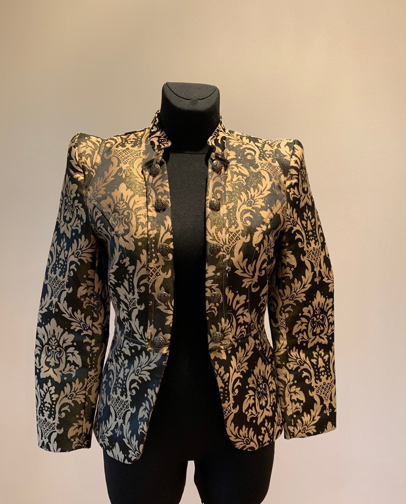 Pode incluir: Um blazer estruturado com um padr&atilde;o de damasco em preto e dourado. O casaco tem gola alta, mangas compridas e um fecho frontal com uma fila vertical de bot&otilde;es met&aacute;licos de cada lado. O tecido apresenta um brilho subtil no design.