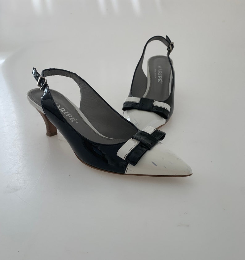 May include: A pair of black and white slingback heels with a bow detail. The heels are made of patent leather and have a pointed toe. The brand name "MARIPE" is printed on the insole.