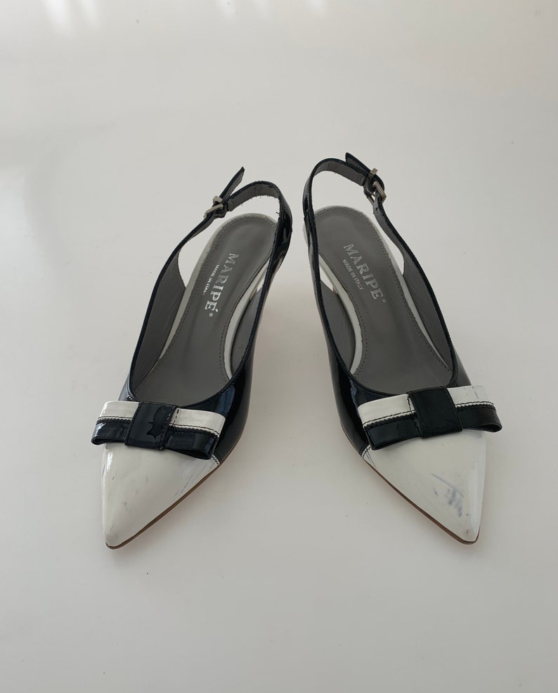 May include: A pair of black and white patent leather slingback heels with a bow detail. The heels are by the brand "MARIPE" and have a small heel.
