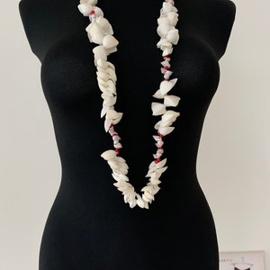 Puede incluir: Un collar de conchas blancas con cuentas rojas. El collar está en un maniquí negro.