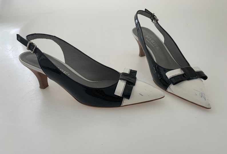 May include: A pair of black and white patent leather slingback heels with a bow detail. The heels are approximately 3 inches tall.
