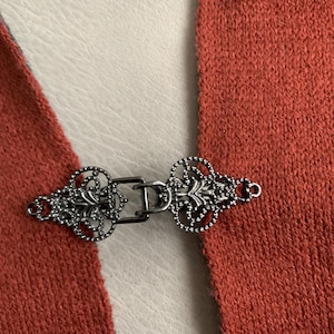 May include: A detailed shot of a decorative metal clasp with an ornate design. The silver-toned clasp has intricate scrollwork and a central bar. It is attached to two sections of rust-coloured knit fabric.
