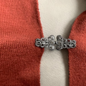 May include: A close-up shot shows a rust-coloured knitted fabric fastened with an ornate silver clasp. The clasp has a detailed, symmetrical design. The fabric appears to be a scarf or wrap.