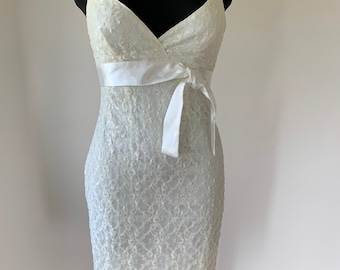 Pode incluir: Um vestido de renda na cor creme, com comprimento até o joelho, alças finas, decote em V e uma fita de cetim lisa amarrada em um laço na cintura. O vestido tem uma silhueta ajustada e é exibido em um manequim preto.