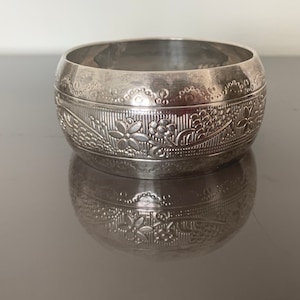 Peut inclure: Un large bracelet jonc en métal argenté orné d'un motif floral en relief et d'une bordure festonnée décorative sur le dessus. La surface présente une finition polie avec des détails complexes qui font tout le tour du bracelet.