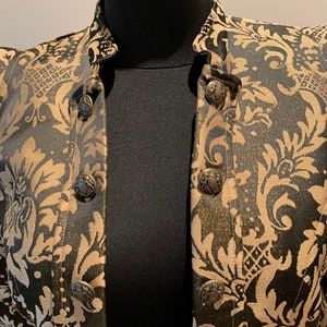 Pode incluir: Um casaco preto e dourado com um padr&atilde;o de damasco proeminente em todo o tecido. A pe&ccedil;a inclui uma gola alta e um fecho frontal fixado por bot&otilde;es met&aacute;licos redondos, gravados com um design de bras&atilde;o detalhado na superf&iacute;cie.