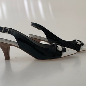 May include: A pair of black and white slingback heels with a bow detail. The heels are made of patent leather and have a wooden heel.