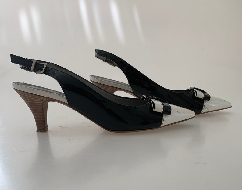 May include: A pair of black and white slingback heels with a bow detail. The heels are made of patent leather and have a wooden heel.