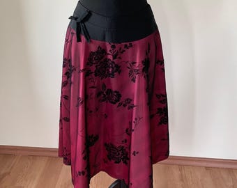 Pode incluir: Uma saia até o joelho com uma base em bordô profundo e um padrão floral de veludo preto. A peça possui um cós largo em preto sólido com um pequeno detalhe de laço decorativo no lado esquerdo. A saia tem uma silhueta evasê em linha A.