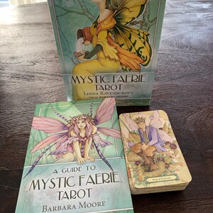 Peut inclure: Le jeu de tarot Mystic Faerie comprend une grande boîte illustrée, un guide intitulé A Guide to Mystic Faerie Tarot par Barbara Moore et un paquet de cartes de tarot avec la carte du Roi de Deniers sur le dessus, orné d'illustrations de fées.