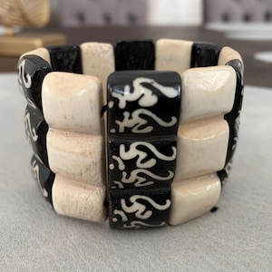 Könnte beinhalten: Ein breites Armband bestehend aus drei Reihen rechteckiger Perlen. Die Perlen wechseln sich zwischen einfarbigem Cremeweiß und Schwarz mit eingravierten weißen Om-Symbolen auf der Oberfläche ab. Das Armband hat eine strukturierte Optik.