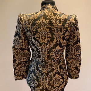 Pode incluir: Um casaco de manga comprida com um padr&atilde;o de damasco em preto e dourado. A pe&ccedil;a tem uma gola alta, ombros estruturados e uma silhueta ajustada. O design floral intrincado cobre toda a superf&iacute;cie do casaco.