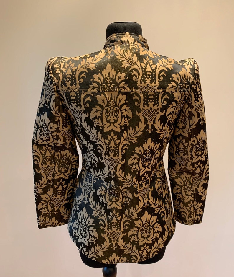 Pode incluir: Um casaco de manga comprida com um padr&atilde;o de damasco em preto e dourado. A pe&ccedil;a tem uma gola alta, ombros estruturados e uma silhueta ajustada. O design floral intrincado cobre toda a superf&iacute;cie do casaco.