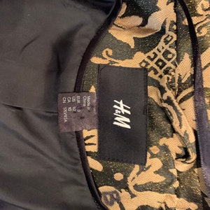 Pode incluir: Uma jaqueta da marca H&M com um padr&atilde;o de damasco em preto e dourado met&aacute;lico. O interior &eacute; forrado com tecido preto s&oacute;lido. Uma etiqueta preta exibe a marca H&M, com uma etiqueta menor indicando Made in China, tamanho US 10 e CN 170/92A.