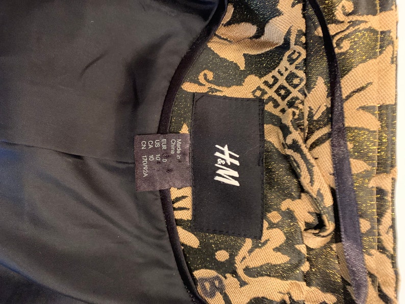 Pode incluir: Uma jaqueta da marca H&M com um padr&atilde;o de damasco em preto e dourado met&aacute;lico. O interior &eacute; forrado com tecido preto s&oacute;lido. Uma etiqueta preta exibe a marca H&M, com uma etiqueta menor indicando Made in China, tamanho US 10 e CN 170/92A.
