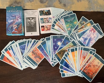 Puede incluir: Una baraja Tarot of Mermaids que incluye una caja azul con la ilustración de una sirena, un folleto de instrucciones y una colección de cartas de tarot extendidas. Las cartas muestran varias ilustraciones acuáticas coloridas y números romanos.